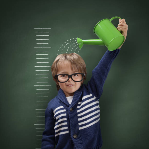 Young boy trying to make himself taller with watering can measuring his growth in height against a blackboard scale